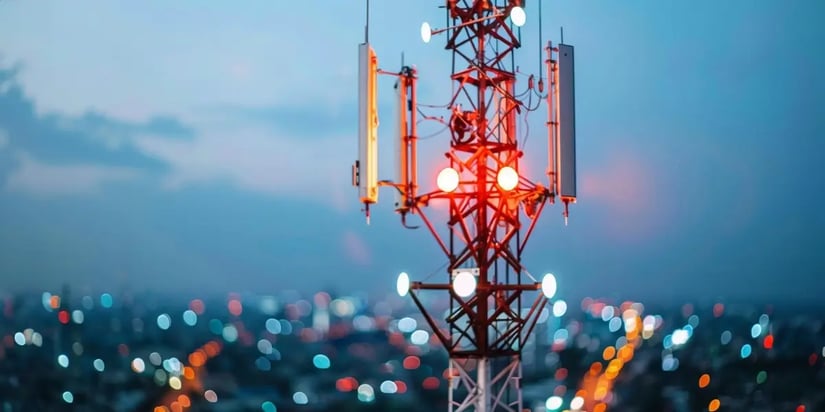 Telecommunication tower illuminated at dusk with city lights in the background.